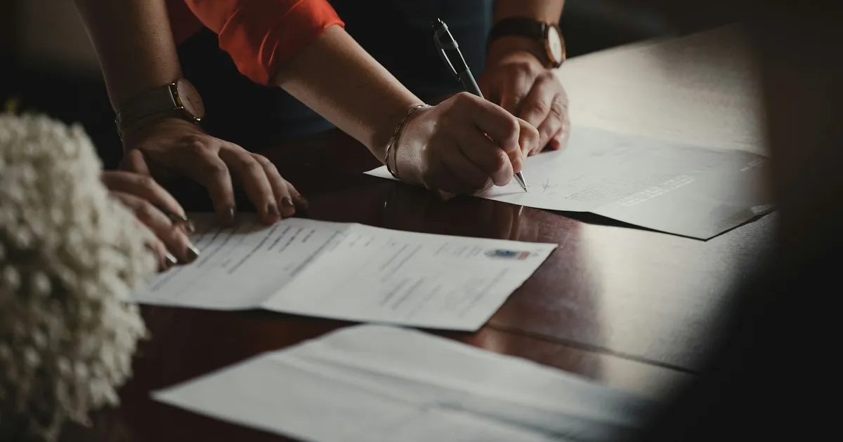 A person in a dark shirt signing a printed document on a wooden desk with a black pen